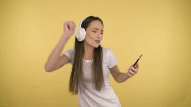 Funny attractive woman 35 years old middle age on yellow background in studio in profile. Woman having fun dancing listening music in big white wireless headphones holding smartphone in her hand