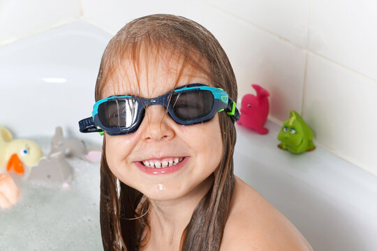 A small, beautiful girl with long hair plays in the bathroom in swimming goggles with rubber toys. The child dives into the bathtub with soap bubbles. Children's hygiene. Water games.