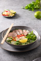 Pho Bo Soup with beef, rice noodles, lime, chili pepper in gray bowl on gray backgound. Vietnamese cuisine. Vertical format.