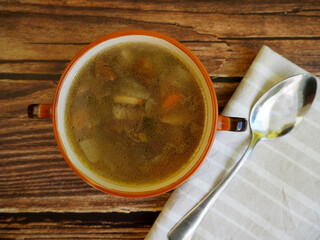 mushroom soup with Polish mushroom, carrots, potatoes and onions in a brown plate with handles stands on a brown wooden table next to a spoon. top view