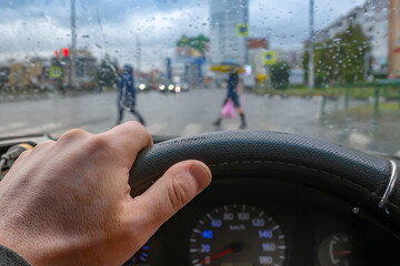 view of the driver hand on the steering wheel of a car against the background of a wet windshield with raindrops and people walking along a pedestrian crossing at an intersection with a traffic light