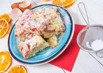 Detail of strawberry souffle cake on blue plate and wheels of dried orange, eggshells on white wooden boards.