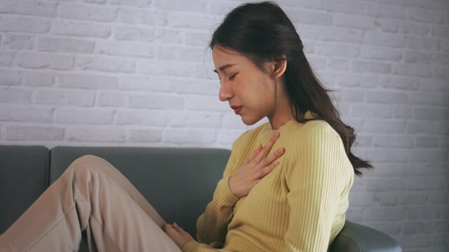 Asian young woman feeling discomfort as suffering from heartburn holding chest with closed eyes and sitting with folded legs on couch at home