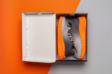 Pair of gray mesh fabric sneakers with grooved orange sole in the open box against orange gray background. New textile trainers for fitness and active lifestyle. Opening new shoes box. Top view.