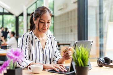 Front view of young Asian woman sitting at office online shopping by paying via credit card in hand with a laptop.