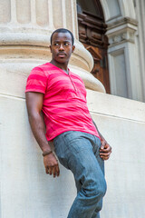 Dressing in red, pink stripe Henley V Neck T shirt,  gray pants, wearing a bracelet, a young black guy is standing against the base of a column  outside an office, waiting for you..