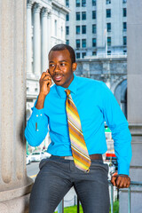 Dressing in a light blue shirt, gray pants,  a pattern tie, a young black businessman is sitting outside an office building, making a phone call..