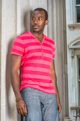 Dressing in red, pink stripe Henley V Neck T shirt,  gray pants, wearing a bracelet, a young black guy is standing by a pillar, confidently looking forward..