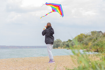 teenage girl launches rainbow kite on river beach. outdoor activity and healthy lifestyle concept.