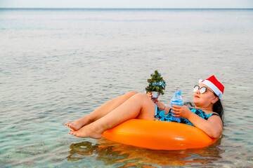 A woman in a swimming ring in a Santa Claus hat with a cocktail and a small Christmas tree in her hand floats in the sea during Christmas holidays.
