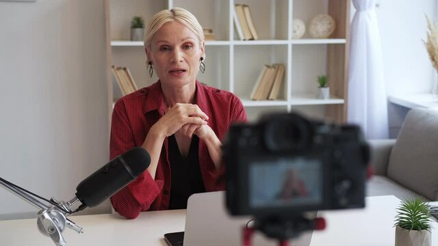 Influencer vlog. Live streaming. Social media communication. Successful mature woman greeting waving hi recording interview on camera on tripod in light home studio interior.