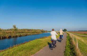 Elderly man and woman couple cycle one behind the other with their electric bicycles on a Dutch dike along the water of an old creek. It is a sunny day in the autumn season.
