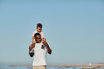 Minimal portrait of happy African-American father carrying son on shoulders against blue sky while...