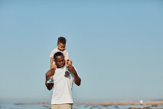 Minimal portrait of happy African-American father carrying son on shoulders against blue sky while enjoying walk on beach in sunlight, copy space