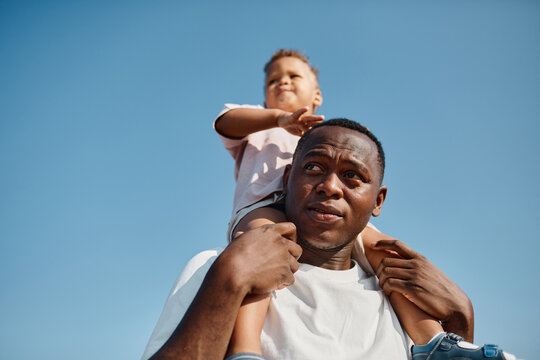 Low angle portrait of happy African-American father carrying son on shoulders against blue sky while enjoying walk in sunlight, copy space