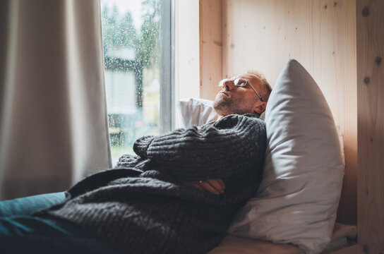 Sleeping Middle-aged man in eyeglasses dressed open cardigan lying on cozy bed pillow next to window. Mental health and autumn weather concept image.