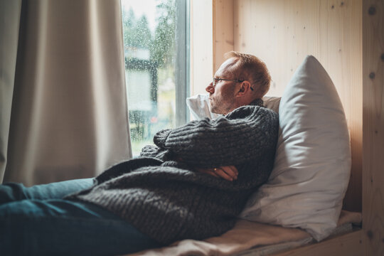 Portrait of thinking sad Middle-aged man in eyeglasses dressed open cardigan lying on cozy bed next to window looking on street through raindrop glass. Mental health and autumn weather concept image.