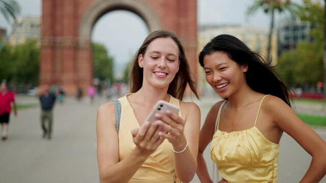 Two happy mixed-race women friends making selfie outdoors and choosing best photo on mobile phone