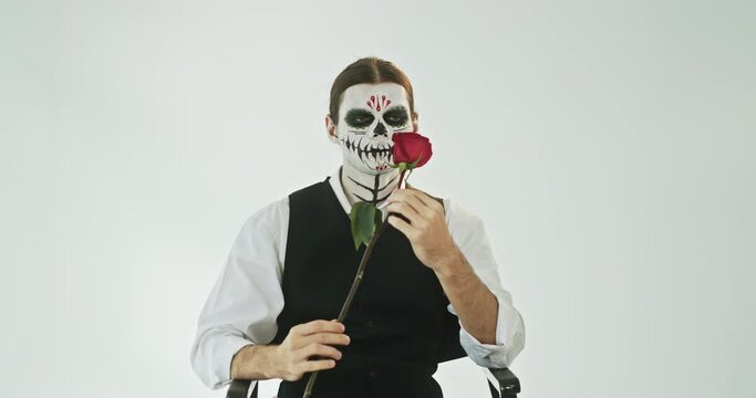The image of a dead romantic on Halloween man in a suit with a rose in his hands looks horrible at the camera sitting on a white background makeup sugar skull