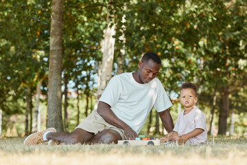 Portrait of young African-American father playing with cute son in park while sitting on grass, copy space