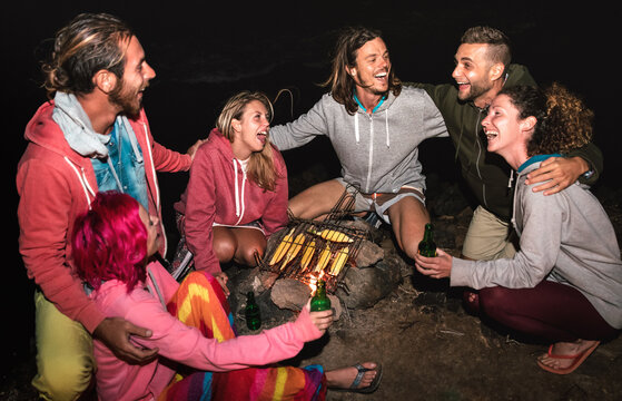 Young indie friends having fun together at night beach party cooking cobs at campfire - Friendship travel concept with alternative people travelers drinking beer at summer bonfire - High iso image