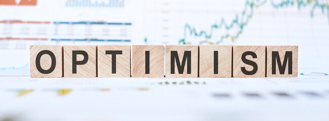 Word OPTIMISM made with wood building blocks on background from financial graphs and charts.