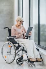 Fototapeta na wymiar Disabled Senior Woman Sitting Near The Window And Using Digital Tablet