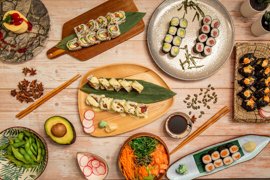 Top view image of Japanese food dishes, salmon maki, uramaki california roll with sesame seeds, edamame, salmon poke bowl, wakame salad, star anise, avocado with nugget and wooden chopsticks.