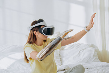 Joyful woman with stylish bionic prosthesis arm and virtual reality goggles plays video game...