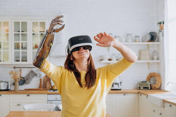 Positive young woman in casual clothes with modern bionic arm and virtual reality headset dances near sofa in stylish kitchen