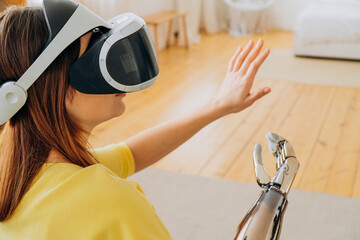 Positive young woman in casual clothes with modern bionic arm and virtual reality headset dances...
