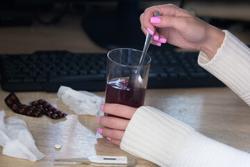 Women's hands on the desktop in front of the computer interfere with the medicine in a glass for colds and pains, next to a computer keyboard, a thermometer, pills, napkins