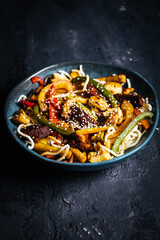 Wok with noodles, peppers, mushrooms, turkey meat and different vegetables and sesame seeds in a blue bowl on dark background