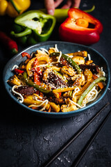Wok with noodles, peppers, mushrooms, turkey meat and different vegetables and sesame seeds in a blue bowl on dark background