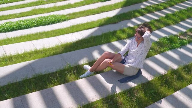 Woman with colorful sunglasses sits on lawn in urban park and makes selfie on smartphone. Summer heat in town.