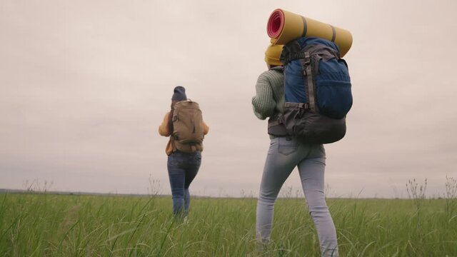 Happy active hipster girls with backpacks on their backs travel through a beautiful valley, a millennial woman on a green field, travel hiking, hiking on the trail, outdoor recreation