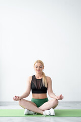 Fototapeta premium Home meditation. Harmony woman. Healthy mind. Yoga balance. Inspired lady in black green sportswear sitting lotus pose closing eyes in light room interior.