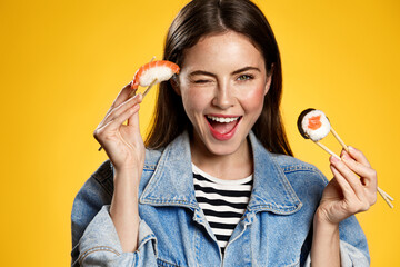 Happy smiling girl eating sushi rolls with chopsticks, holding sashimi and winking at camera, order...