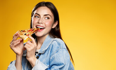 Photo of attractive woman eats slice of pizza, points aside with thumb, dressed in fashionable clothes, shows where pizzeria is, isolated over yellow background. Pretty girl has snack with fastfood - obrazy, fototapety, plakaty