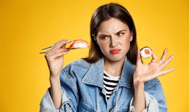 Picky Girl Looking At Sashimi With Disgust, Dont Like Raw Fish Rolls In Japanese Restaurant, Standing Displeased Against Yellow Background