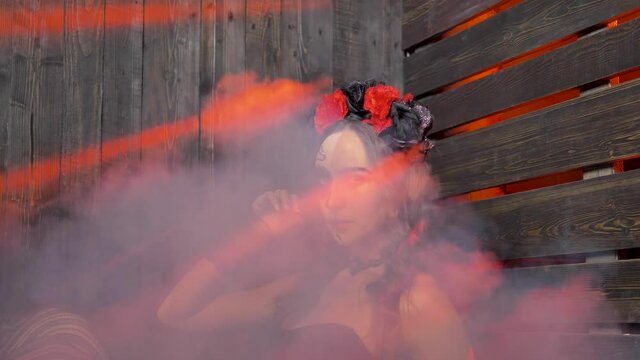 A portrait of a lovely woman with Halloween makeup on her face in red rays of light is enveloped in clouds of white smoke. He looks intently at the camera.
