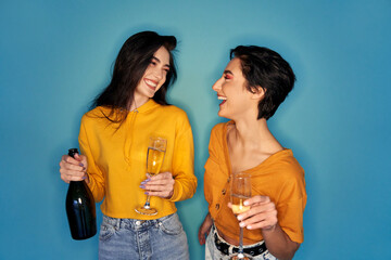 Two happy pretty stylish cool diverse gen z girls friends having fun celebrating night club hen party, dancing, holding glasses, laughing and drinking champagne on green blue studio background. - obrazy, fototapety, plakaty