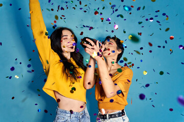 Two happy excited pretty stylish cool diverse gen z girls friends having fun celebrating New Year or birthday hen night club party event, dancing in confetti and drinking champagne on blue background. - obrazy, fototapety, plakaty