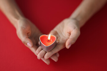 Red burning candle in the shape of a heart in female hands on red background