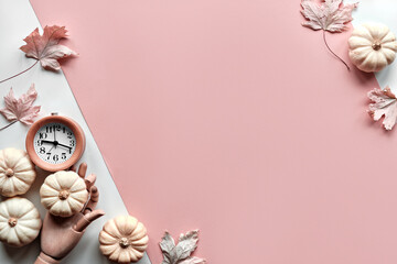 Autumntime background. Alarm clock in wooden model hand, and decorative white pumpkins. Layered off white and orange paper flat lay with dry maple leaves. Seasonal arrangement, monochromatic look.