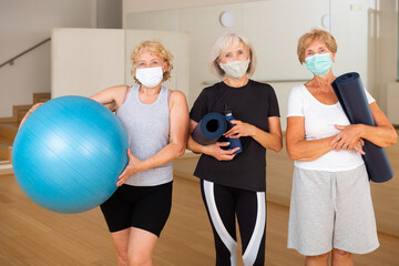 Three smiling aged women in sportswear and protective masks standing in fitness studio with sports...