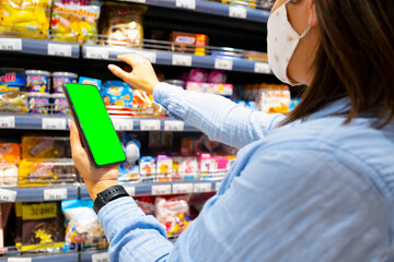 Cropped photo of a young woman with face mask using mobile phone and buying groceries in the supermarket during virus pandemic.