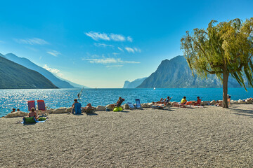 Fototapeta na wymiar Torbole, Italy-09 October 2018: People sitting at the beach on the lounge chair and admiring Lake Garda in the summer time,View of the beautiful Lake Garda surrounded by mountains