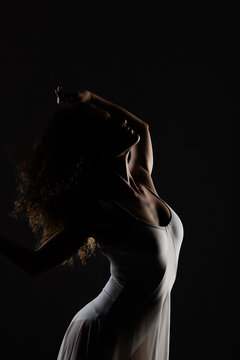 Girl with curly hair making ballet poses. Side lit silhouette of ballerina in white dress against black background.