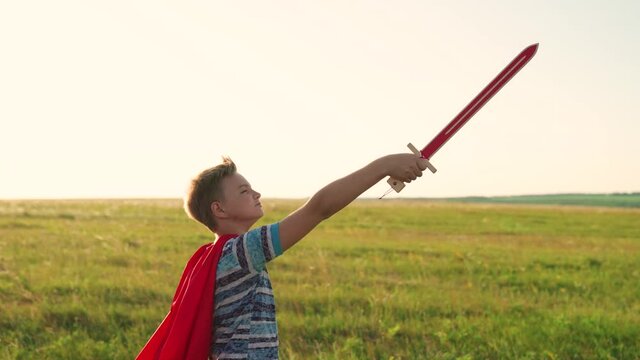 A free boy in a red cloak with a sword in his hand plays a medieval knight in sun. Boy fights with a toy sword. Child plays superheroes. Teenager plays the Spartan. Happy childhood concept.